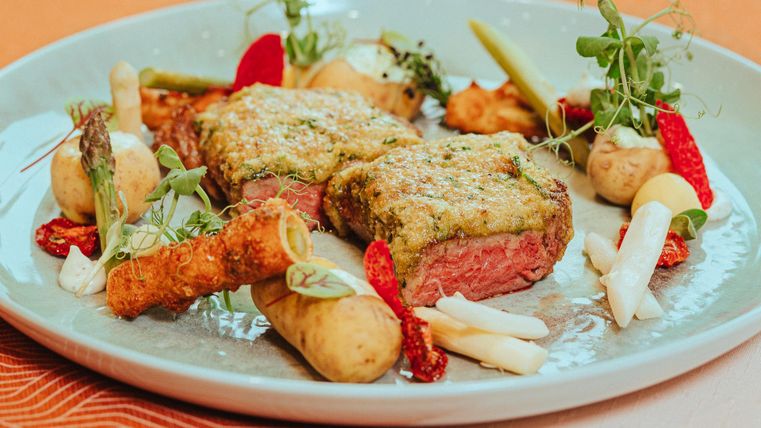 Een heerlijk gerecht met twee stukken vlees, bedekt met een kruidenkorst. Omringd door verschillende groente bijgerechten en verse kruiden.