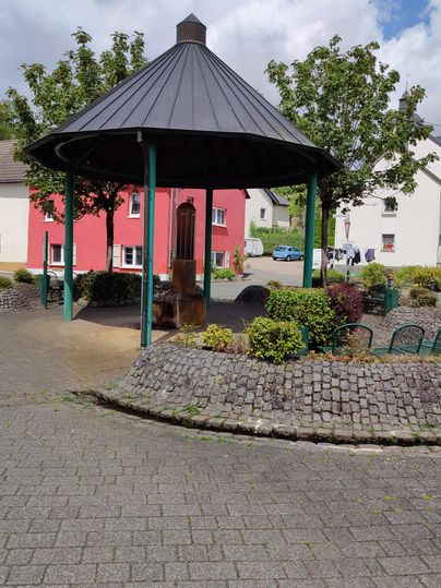 A pavilion in a small park, surrounded by trees and shrubs. In the background, colorful houses can be seen.