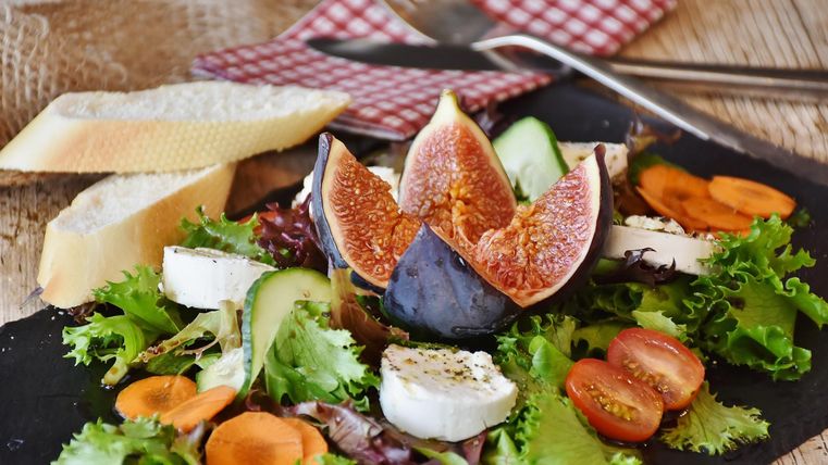 Salad plate topped with green lettuce, tomatoes, cucumber slices, fig, carrot slices, and round cheese slices. At the edge of the square black plate are two slices of baguette.
Symbol image