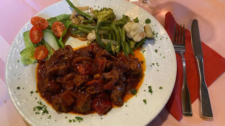 Ein Teller mit herzhaftem Fleischgericht in Sauce, begleitet von frischem Gemüse und Salat. Daneben stehen Besteck und ein Gläschen Wein.