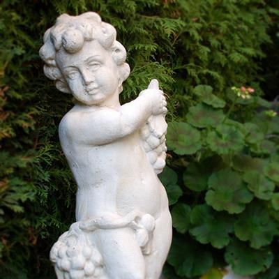 Eine weiße Statue eines Jungen mit lockigem Haar, der Trauben hält. Im Hintergrund sind grüne Pflanzen sichtbar.
