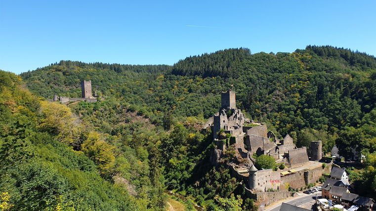 Eine malerische Aussicht auf eine alte Burg inmitten grüner Hügel. Im Hintergrund sind weitere Burgen und ein klarer Himmel sichtbar.