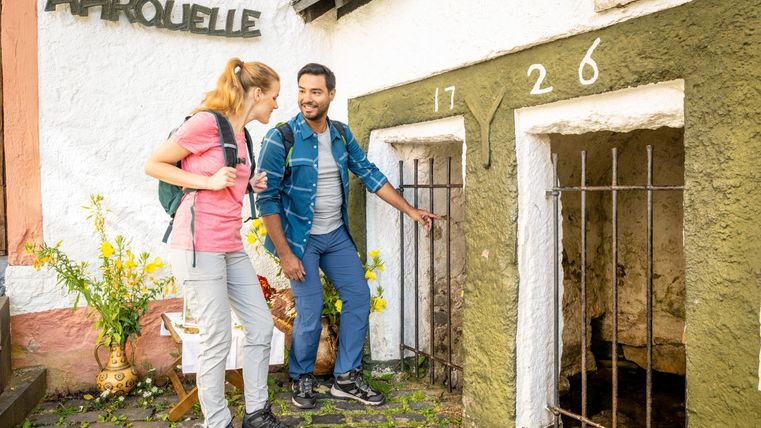 Twee wandelaars staan voor een historisch gebouw met de inscriptie „AHROUELL“. Ze verkennen de ingangen, terwijl om hen heen bloemen bloeien.