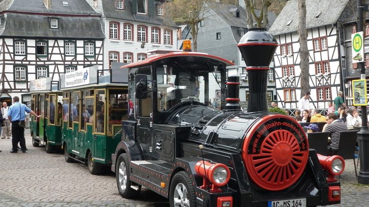 A small train with a black locomotive and red accents is parked in a picturesque town with timber-framed houses. The street is paved, and some people can be seen nearby.