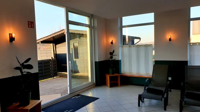 Een lichte kamer met grote ramen en uitzicht op de buitenruimte. Er zijn comfortabele ligbedden en enkele planten ter decoratie.