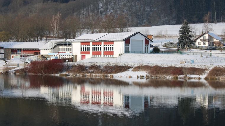 A modern building by the shore of a tranquil lake, surrounded by snow. The landscape is clear and reflected in the water.