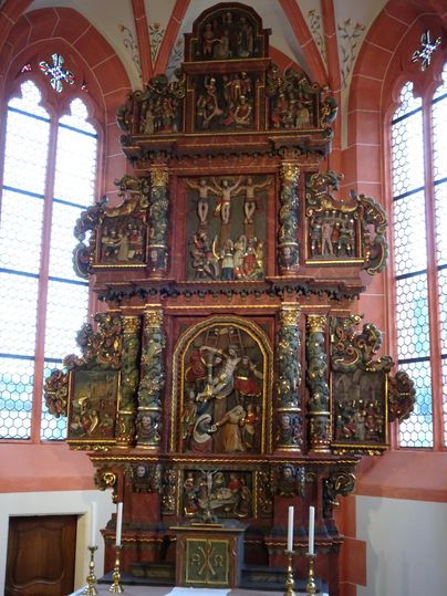 Een kunstzinnig gesneden altaar van hout, versierd met religieuze figuren. Het altaar staat in een kerk en heeft grote ramen die licht binnenlaten.