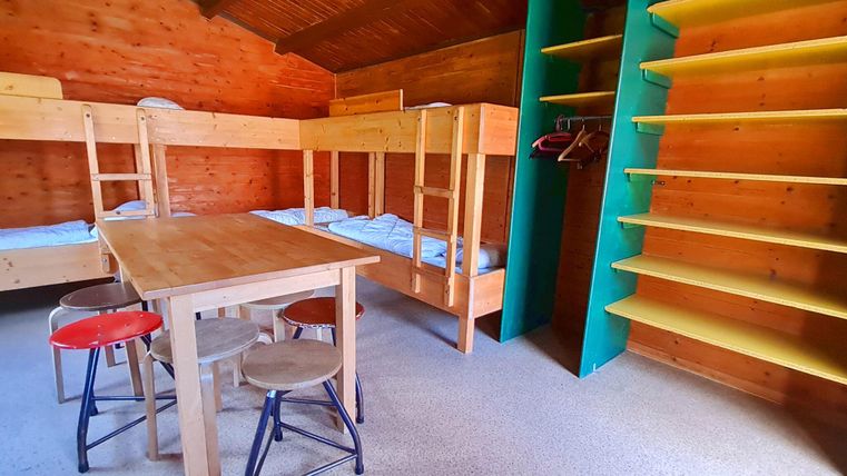 Ein einfaches Zimmer mit zwei Etagenbetten und einem Tisch in der Mitte. An der Wand befindet sich ein Regal zur Aufbewahrung.