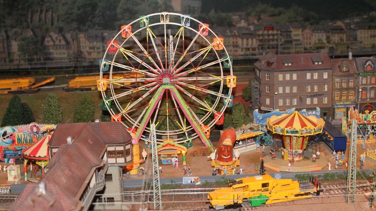 A small fairground model with a ferris wheel and a carousel. In the background, buildings and trains are visible.