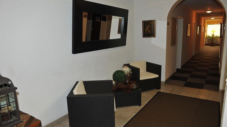 A bright hallway with white walls and a dark carpet. There are seating areas made of rattan and a large mirror on the wall.