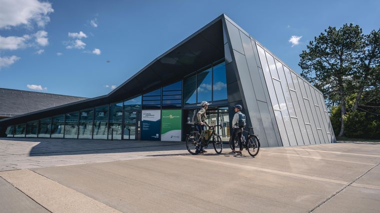 Ein modernes Gebäude mit einer großen Glasfront und einer asymmetrischen Dachform. Zwei Fahrradfahrer stehen vor dem Eingang und genießen den sonnigen Tag.