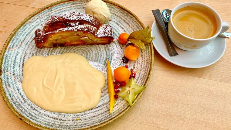 A plate with apple strudel, cream, and colorful fruit. There is also a cup of coffee in a light saucer.