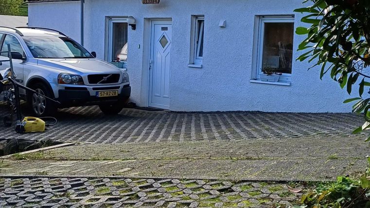 Ein kleines, weißes Haus mit einem grauen Dach steht auf einem gepflasterten Vorplatz. Es sind zwei Autos geparkt und der Himmel ist bewölkt.