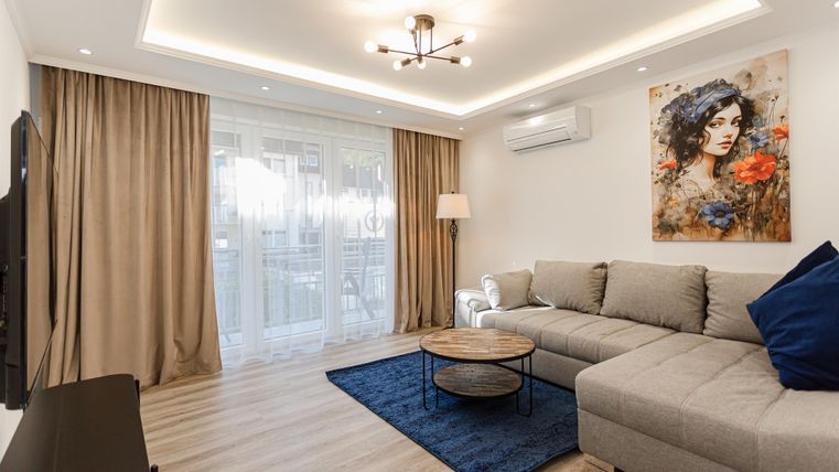 Ein modernes Wohnzimmer mit einem grauen Sofa und einem runden Tisch. An der Wand hängt ein farbenfrohes Kunstwerk und große Fenster sorgen für viel Tageslicht.