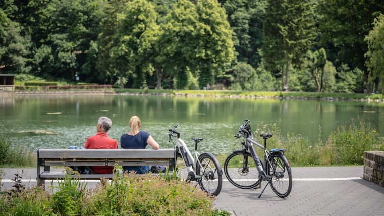 Een paar zit op een bank aan de oever van een meer. Naast hen staan fietsen en bomen omgeven het vredige tafereel.