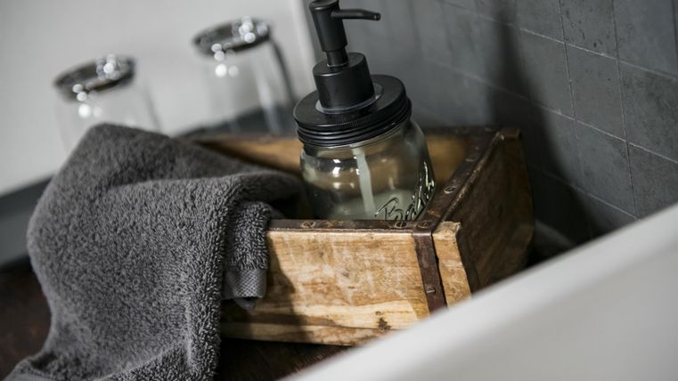 Eine Holzbox mit einem Seifenspender und einem Handtuch liegt am Rand eines Waschbeckens. Die Atmosphäre ist schlicht und modern.