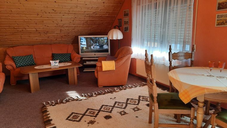 A cozy living room with orange walls and a wooden floor. The furnishings include a seating area with couches, a table, and a television.