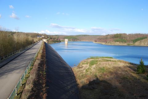 Ein ruhiger Stausee umgeben von Wäldern und Hügeln. Eine asphaltierte Straße verläuft entlang des Ufers.
