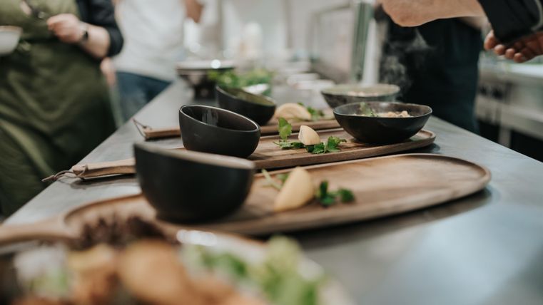 Eine moderne Küche mit verschiedenen Schalen und frischen Zutaten auf einem Holzbrett. Köche arbeiten im Hintergrund an der Zubereitung von Gerichten.