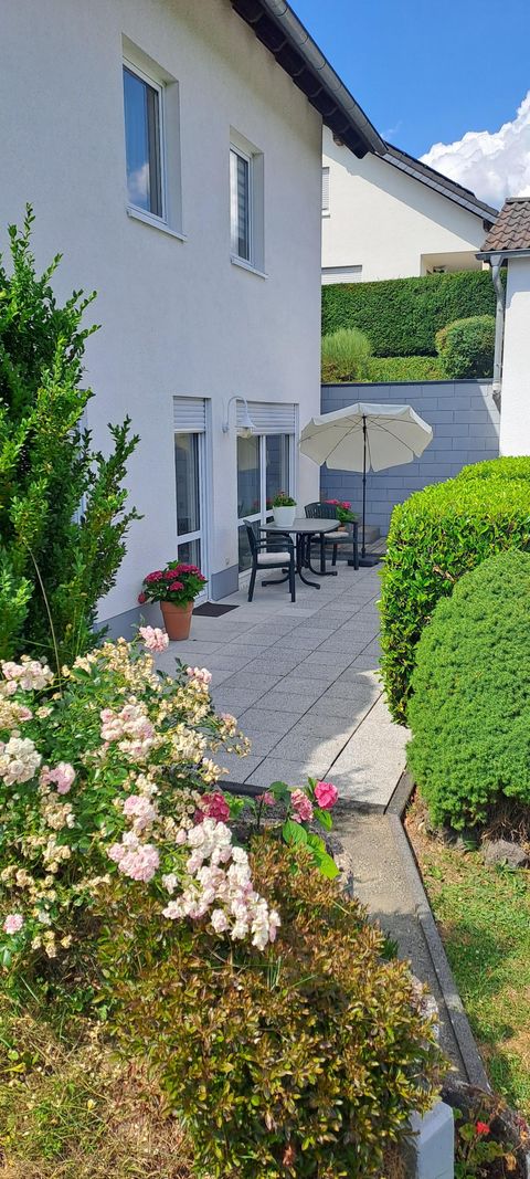Een gezellige terras met bloemen en een parasol. Op de achtergrond is een wit huis en goed onderhouden hagen te zien.