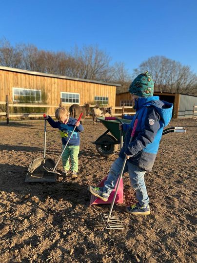 Zwei Kinder spielen auf einem Bauernhof und nutzen Werkzeuge, um den Boden zu bearbeiten. Im Hintergrund sind ein Stall und einige Bäume sichtbar.