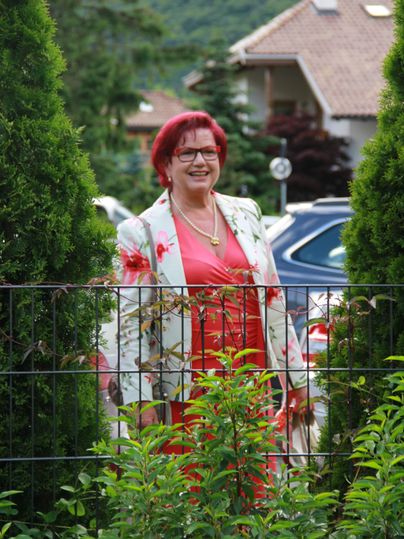 Eine Frau mit roten Haaren steht vor einem Zaun, umgeben von grünen Pflanzen. Sie trägt ein blumiges Jackett und lächelt freundlich.