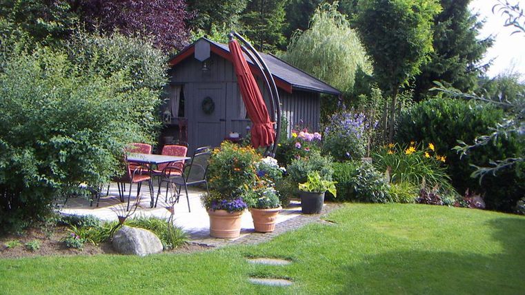 A lovely garden with colorful flowers and a cozy seating area. In the background, there is a small garden shed surrounded by trees.