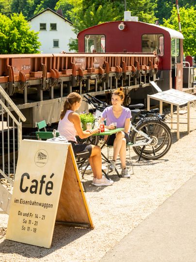 Een sfeervol café in een spoorwegwagon met twee vrouwen die aan tafel zitten. Fietsen staan ernaast, omringd door groene natuur.