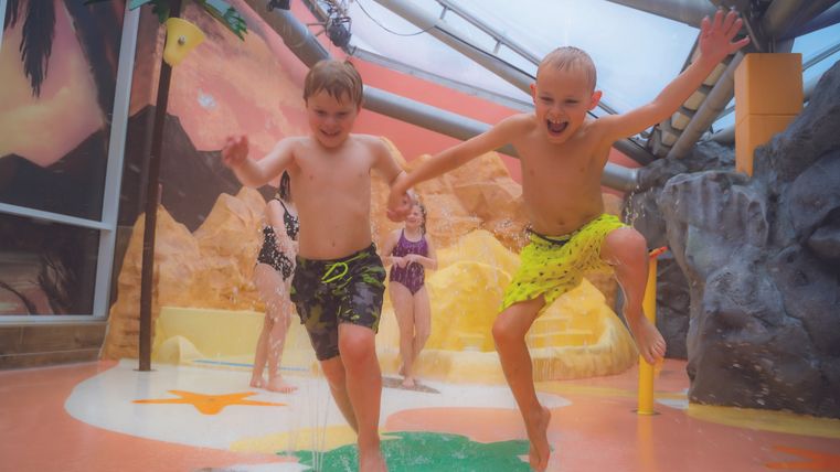 Zwei fröhliche Kinder springen in einem Wasserpark. Im Hintergrund sind bunte Spielgeräte und eine entspannte Urlaubsatmosphäre zu sehen.