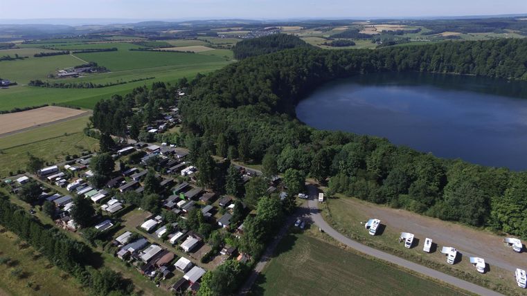 Ein malerischer Campingplatz am Ufer eines Sees, umgeben von üppigem Grün und Feldern. Die ruhige Landschaft lädt zur Erholung in der Natur ein.