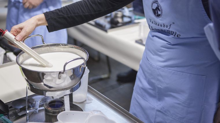 Eine Person in einer blauen Schürze bereitet in einer Küche Milch zu. Im Hintergrund sind weitere Kochutensilien sichtbar.