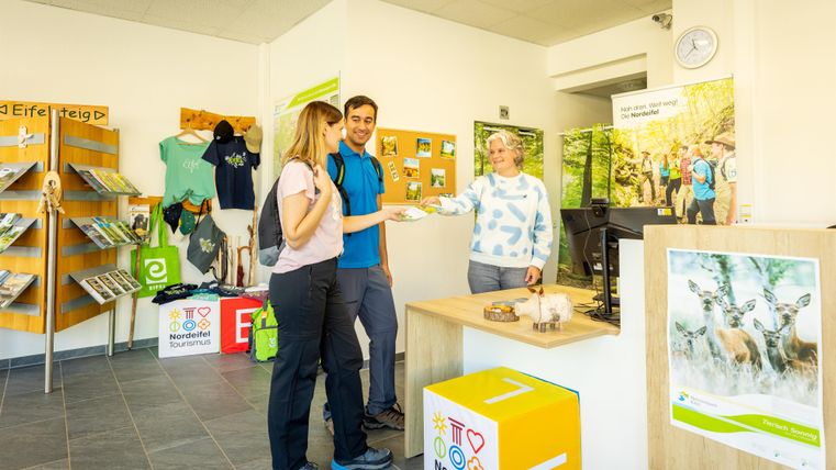 Ein freundlicher Empfangsbereich mit zwei Gästen und einer Mitarbeiterin. Es gibt Broschüren und Merchandise-Artikel für Wanderer.