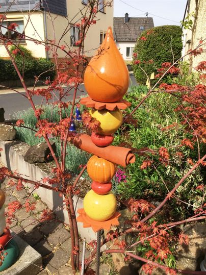 Een kleurige tuin decoratiepaal met verschillende vormen in oranje en geel. Op de achtergrond zijn herfstachtige takken en een rustige straatzicht te zien.