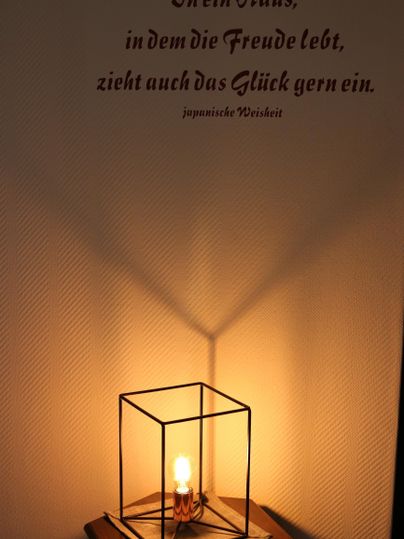 Eine stilvolle Lampe in einem geometrischen Gestell steht auf einem Tisch. An der Wand hängt ein inspirierendes Zitat über Freude und Glück.