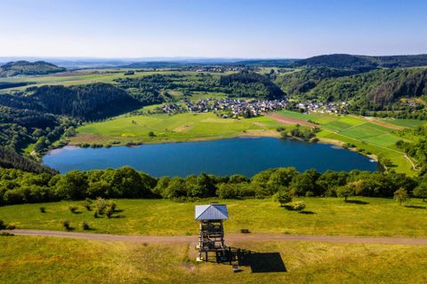 Eine malerische Landschaft mit einem See, umgeben von grünen Wiesen und Hügeln. Im Hintergrund ist ein kleines Dorf zu sehen.
