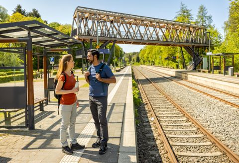Ein Bahnhof im Freien mit zwei Reisenden, die miteinander sprechen. Im Hintergrund sind Gleise und ein Fußgängerüberweg zu sehen.
