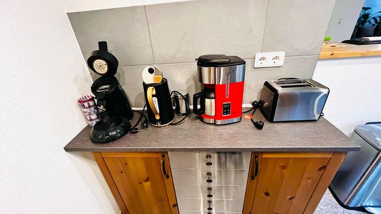 A modern kitchen unit with various appliances, including a coffee machine, a kettle, a toaster, and a kitchen helper. The room is bright and friendly designed.
