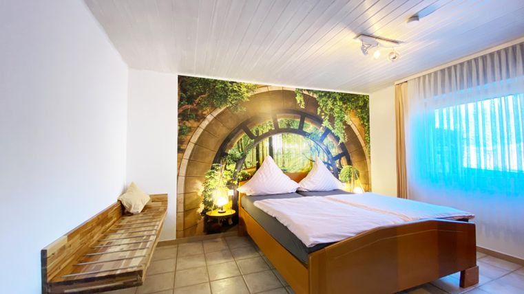 Ein gemütliches Schlafzimmer mit einem großen Bett und einer Holzbank. Im Hintergrund befindet sich eine Wand mit einem grünen Motivelement, das eine natürliche Atmosphäre schafft.