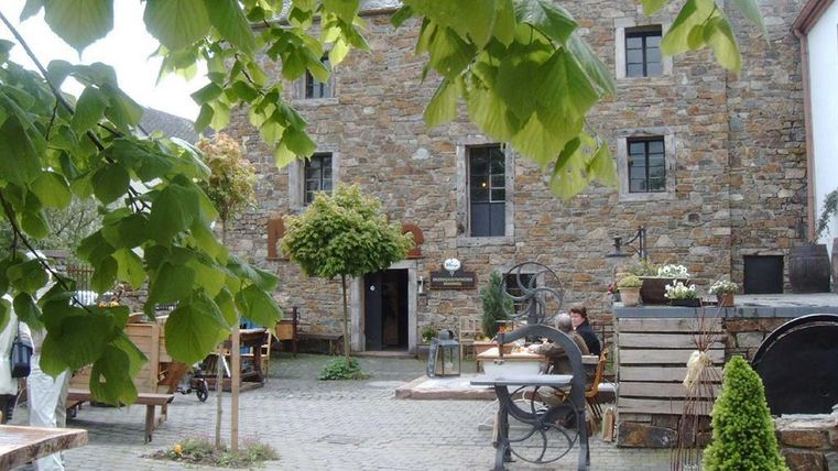 Courtyard of a historic mill with stone walls, tables, and plants. People are sitting at tables, surrounded by green leaves and rustic decorations.