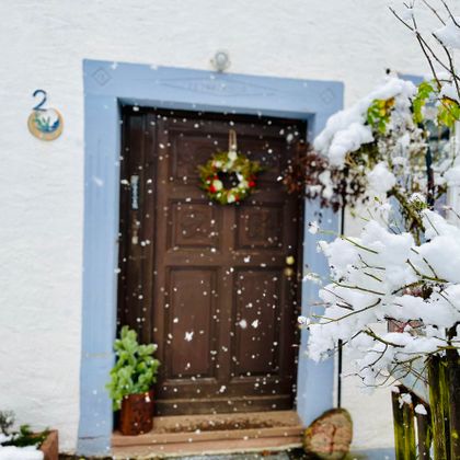 Eine braune Eingangstür mit einem runden Türkranz ist von Schnee umgeben. Im Vordergrund sind schneebedeckte Pflanzen zu sehen.