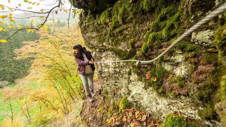 Person wandert entlang eines felsigen, mit Moos bedeckten Pfades, hält sich an einem Seil fest. Herbstliche Bäume im Hintergrund.