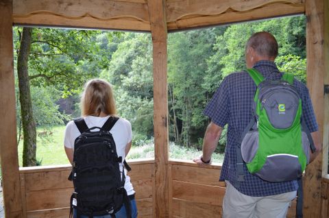 Zwei Wanderer stehen in einem Holzsichtschutz und schauen in die grüne Natur. Beide tragen Rucksäcke und genießen die Aussicht.