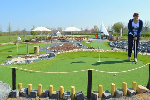 Eine Frau spielt Mini-Golf auf einem schön gestalteten Platz mit grünem Rasen und Steinen. Im Hintergrund sind einige Pavillons und Bäume zu sehen.