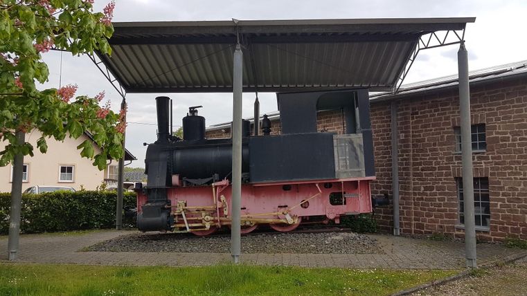 Eine historische Dampflok steht unter einem Vordach. Die Lok ist schwarz mit pinken Akzenten und befindet sich in einem gepflegten Außenbereich.