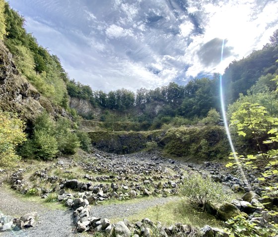 Innenbereich des Arensberg Vulkans mit steinigen Pfaden, umgeben von gr&uuml;ner Vegetation und Felsen unter einem bew&ouml;lkten Himmel mit Sonnenstrahlen., &copy; Touristik GmbH Gerolsteiner Land