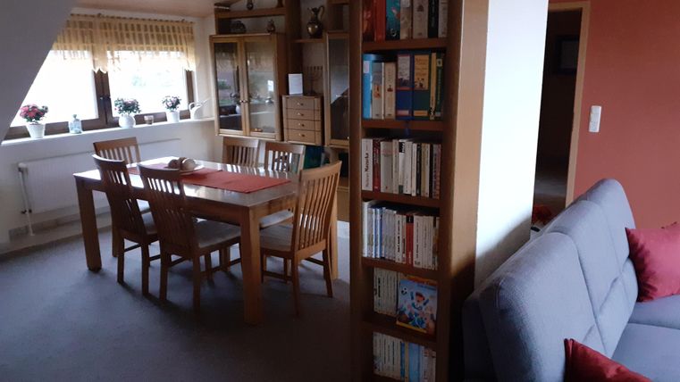 Een gezellige woonruimte met een eettafel en stoelen. Rechts staat een boekenkast vol boeken en een grijze bank.