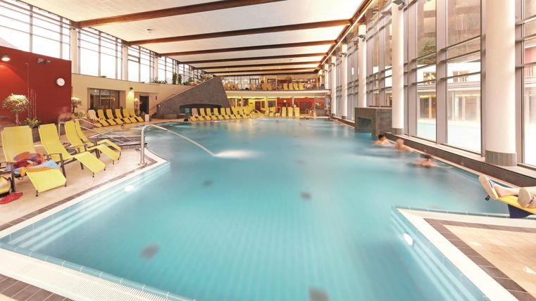 Ein großer Innenpool mit klarblauem Wasser und komfortablen Liegen. Helle Fenster sorgen für eine angenehme Atmosphäre und viel Licht.