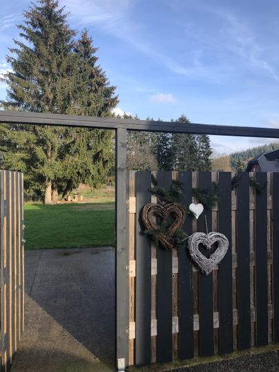 Ein Tor aus Holz mit zwei dekorativen Herzen hängt daran. Im Hintergrund ist ein grüner Garten und ein großer Baum zu sehen.