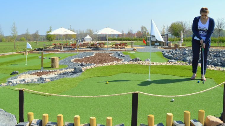 Eine Frau spielt Mini-Golf auf einem schön gestalteten Platz mit grünem Rasen und Steinen. Im Hintergrund sind einige Pavillons und Bäume zu sehen.