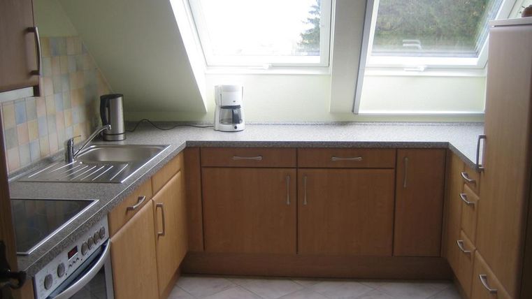 Een moderne keuken met houten kasten en een lichte werkblad. Grote ramen laten veel licht binnen.